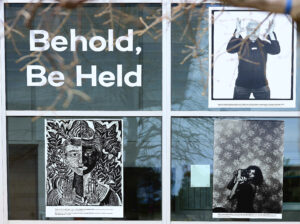 Photograph of Gallery 4 paned window with reflections. Upper left Beheld, Be Hold title; Upper right phto of man holding arms up; lower left Margaret Burroughs Two Worlds,wood block black and white print; lower right Ming Smith, untitled self portrait with camera